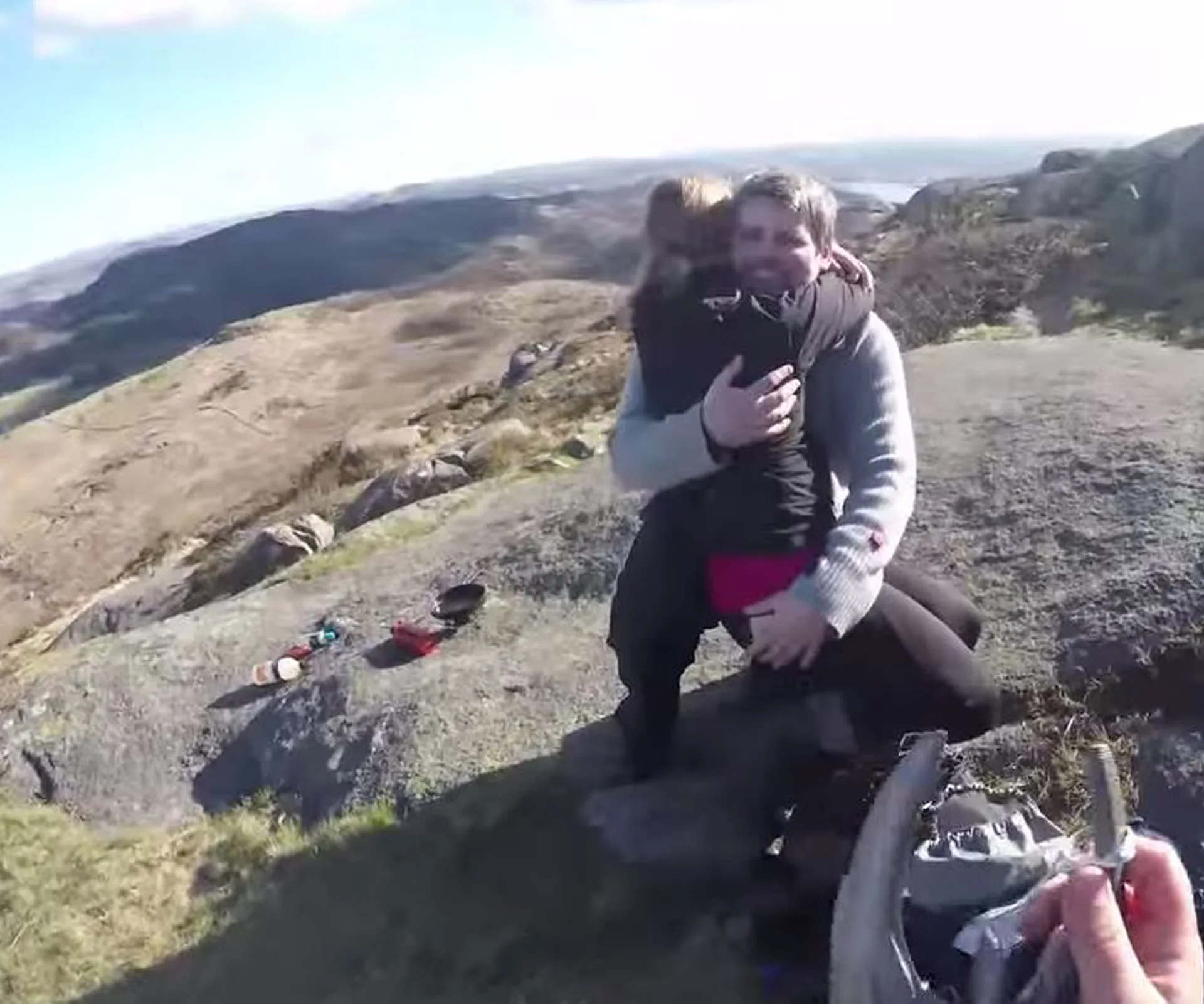 Man kneels on rocky hilltop hugging a woman, with scattered items nearby and scenic landscape in the background.