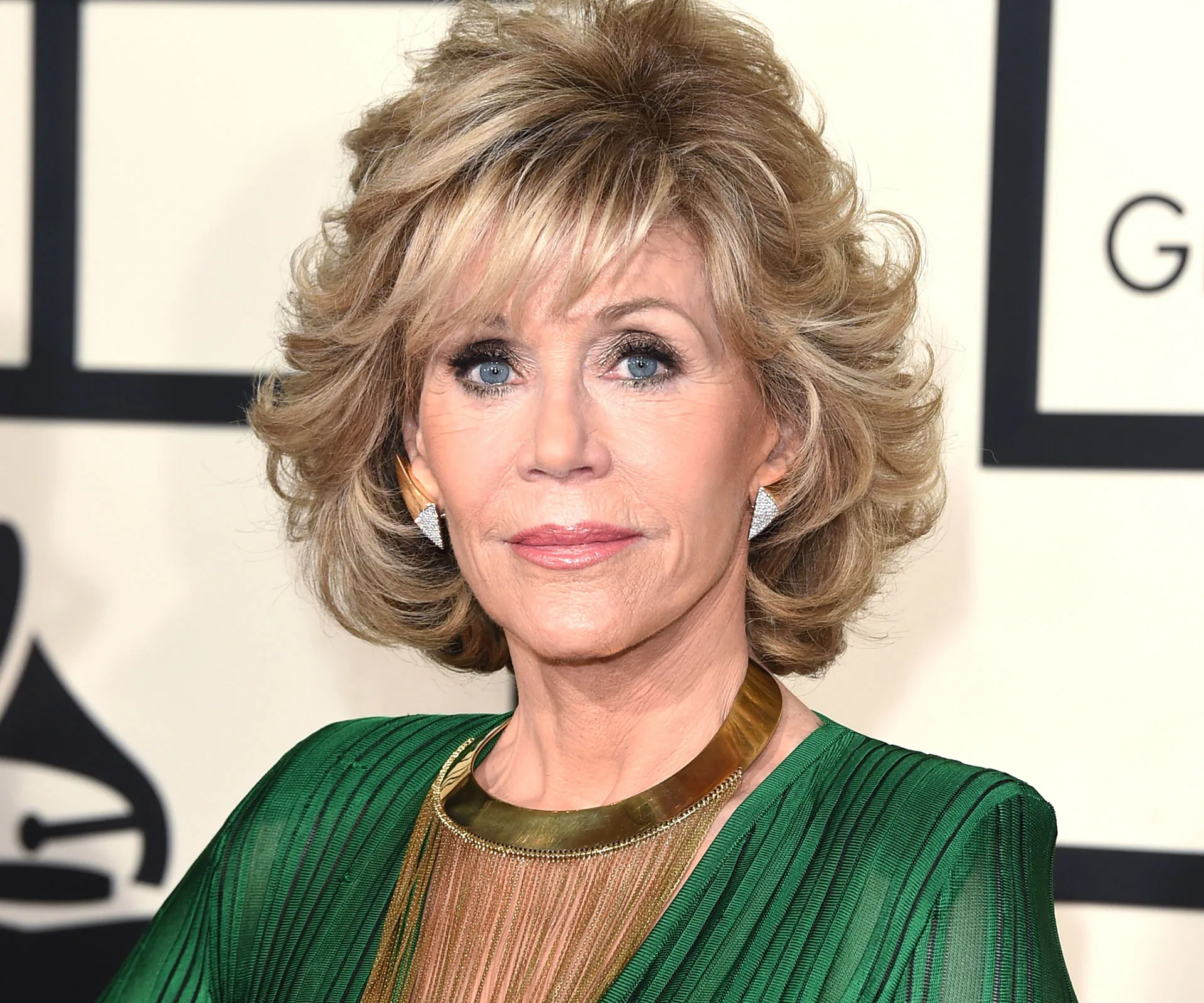 A person with short, styled blonde hair wears a green dress and gold necklace at a formal event.