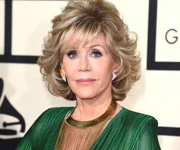 A person with short, styled blonde hair wears a green dress and gold necklace at a formal event.