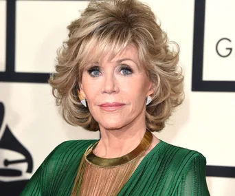 A person with short, styled blonde hair wears a green dress and gold necklace at a formal event.