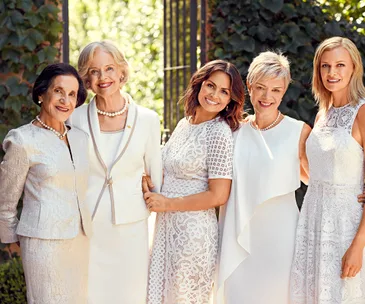 Five women in elegant white dresses and pearls stand together smiling, with greenery in the background.