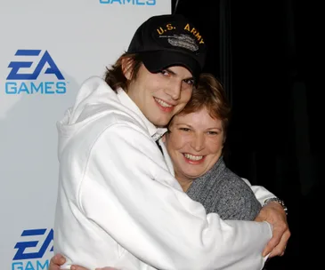 Man in a U.S. Army cap hugging a woman with gray sweater, both smiling, in front of an EA Games backdrop.