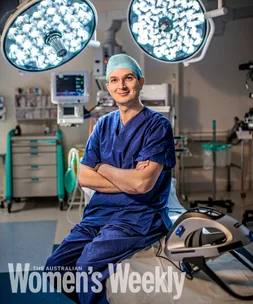Surgeon in scrubs sits on an operating table with arms crossed, surrounded by medical equipment and bright surgical lights.