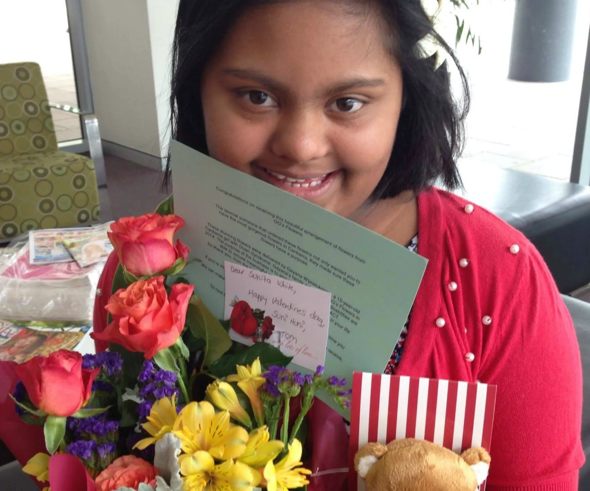 Girl smiling with colorful bouquet and greeting card, dressed in a red cardigan with pearl embellishments.