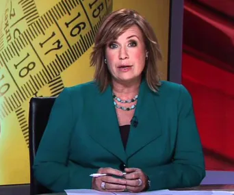 News anchor sitting at a desk with a measuring tape graphic behind, wearing a green blazer and holding a pen.