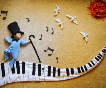 Baby sleeping in blue onesie with top hat on a piano key blanket, surrounded by musical notes and birds.