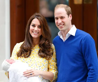 A smiling woman in a yellow floral dress holds a newborn, standing next to a man in a blue sweater outside.