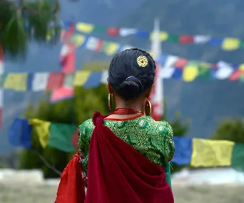 Nepalese woman in traditional attire, facing colorful prayer flags against a mountain backdrop.