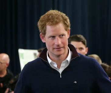 Prince Harry in a navy sweater, surrounded by people at an indoor event.