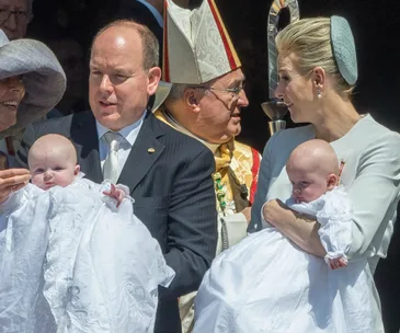 A family of four with two infants in white christening gowns, an adult holding each child, outside a church.