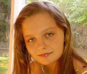 Young girl with light brown hair and a piercing on her lower lip, sitting indoors with natural light from a window behind her.