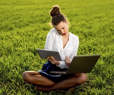 Young woman multitasking with laptop, tablet, and phone, sitting on grass.