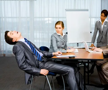 Business meeting; man in suit sleeping in chair, two women and one man working around a table with laptop and notepad.