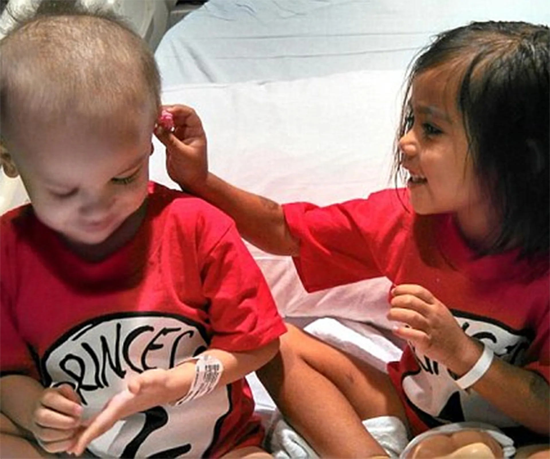 Two young children with short hair, wearing matching red "Princess 1" and "Princess 2" shirts, sit together smiling.