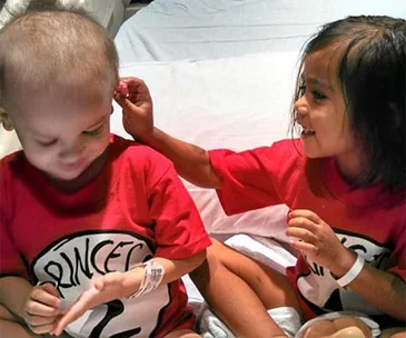 Two young children with short hair, wearing matching red "Princess 1" and "Princess 2" shirts, sit together smiling.
