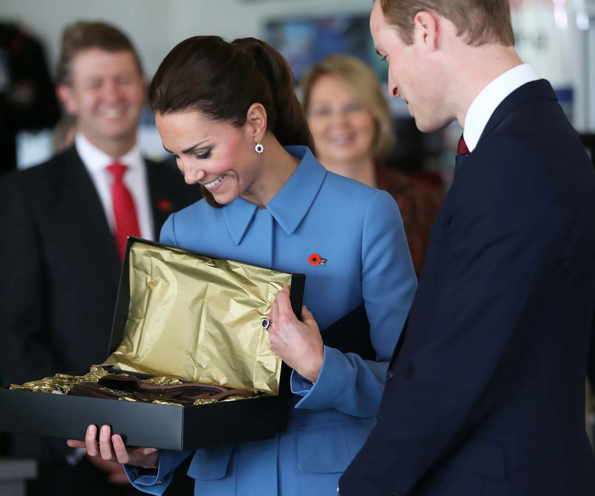 "Person in blue coat smiling at a gift box with gold lining, surrounded by people in formal attire."