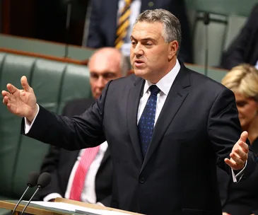 A person in a suit delivers a speech in a formal setting, gesturing with both hands amidst seated colleagues.