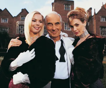 Three elegantly dressed people pose together in front of a large brick mansion.