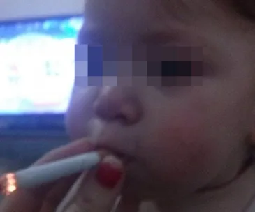 Baby with face blurred, held by a hand with red nails, holding a lit cigarette near the mouth.