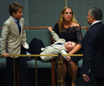 A woman and two boys, dressed formally, sit in a governmental chamber, engaging with a man in a suit.