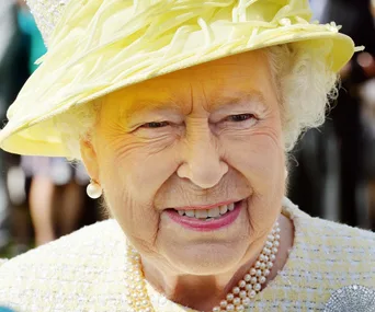 A smiling elderly woman in a yellow hat and pearls.