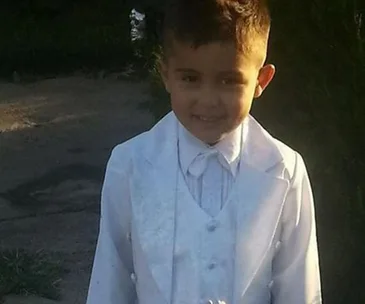 A young boy in a white suit smiles at the camera outdoors.