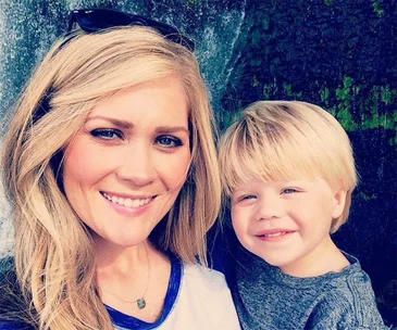 Smiling woman with sunglasses on head embracing smiling blond child in front of a green, textured background.