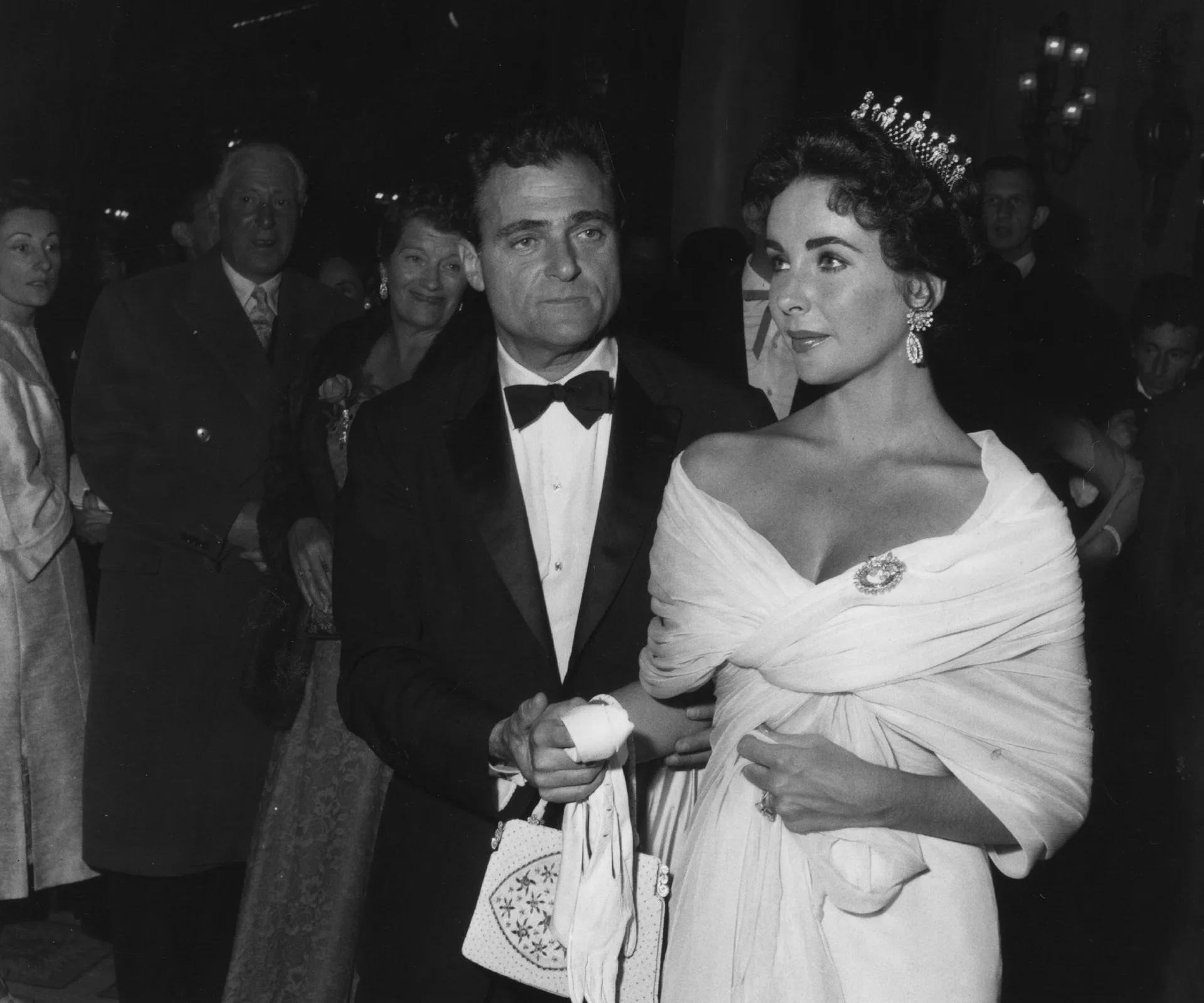 Couple in formal attire at an event; woman in a tiara and elegant gown, holding man's arm.