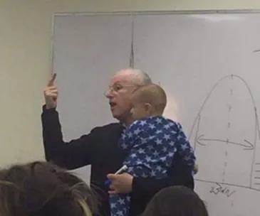 Professor holds a baby in star pajamas while teaching at a whiteboard with a graph.