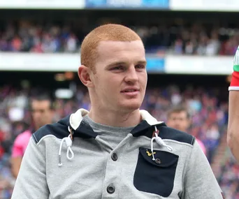 A man in a hoodie sits in a stadium, looking slightly to the side, with a crowd in the background.