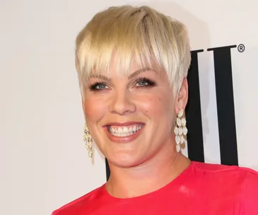 Smiling person with short blonde hair and dangling earrings in a bright pink top.