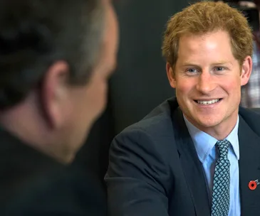 "Smiling man in a suit with light hair, wearing a poppy pin, in conversation with another person."