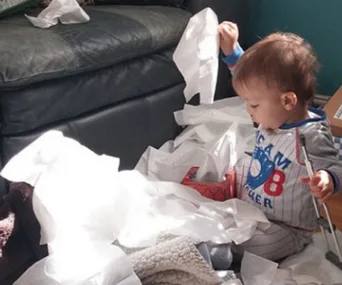 A toddler sitting on a couch surrounded by tissues, playing with them.