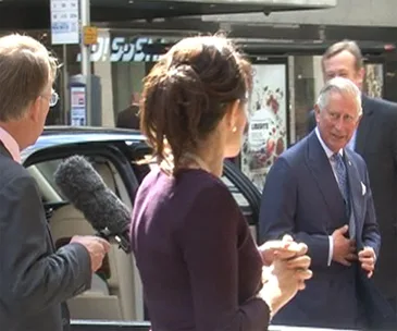 Prince Charles in conversation with reporters outside, one holding a microphone, with a car and building in the background.