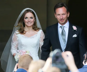 Bride in lace gown and groom in suit smiling, surrounded by photographers.