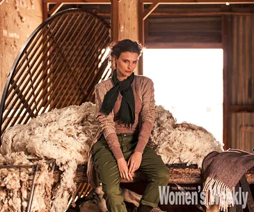 A woman in a rustic setting sits on a bench with wool around, wearing a scarf and sweater, light filtering through a window.