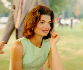 Woman with brown hair in a green dress, smiling and seated outdoors in a park setting.