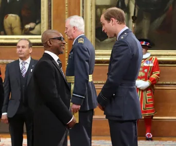 Prince William greets a man in a formal setting, with others in ceremonial attire in the background.