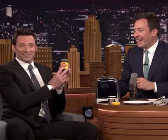 Hugh Jackman shows a jar of Vegemite to Jimmy Fallon on a talk show set, with cityscape backdrop.