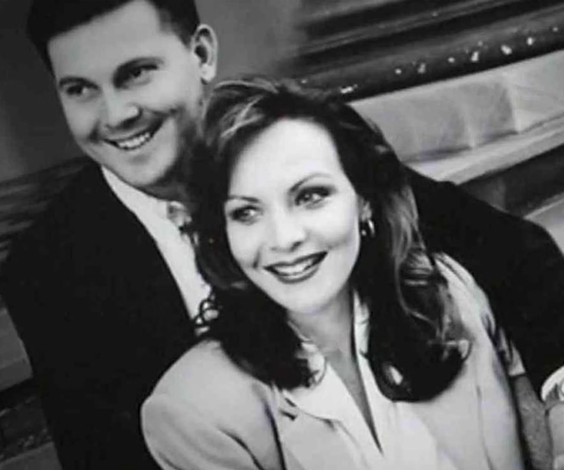 A smiling couple poses together, dressed formally, in a black and white photo.