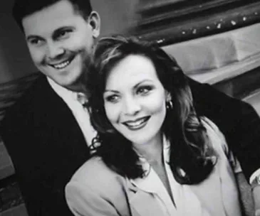 A smiling couple poses together, dressed formally, in a black and white photo.
