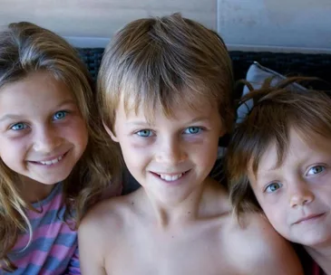 Three smiling children with blue eyes and light brown hair, sitting closely together.