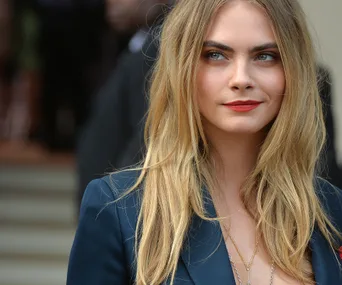 A person with long blonde hair, bold eyebrows, and red lipstick wearing a navy outfit.