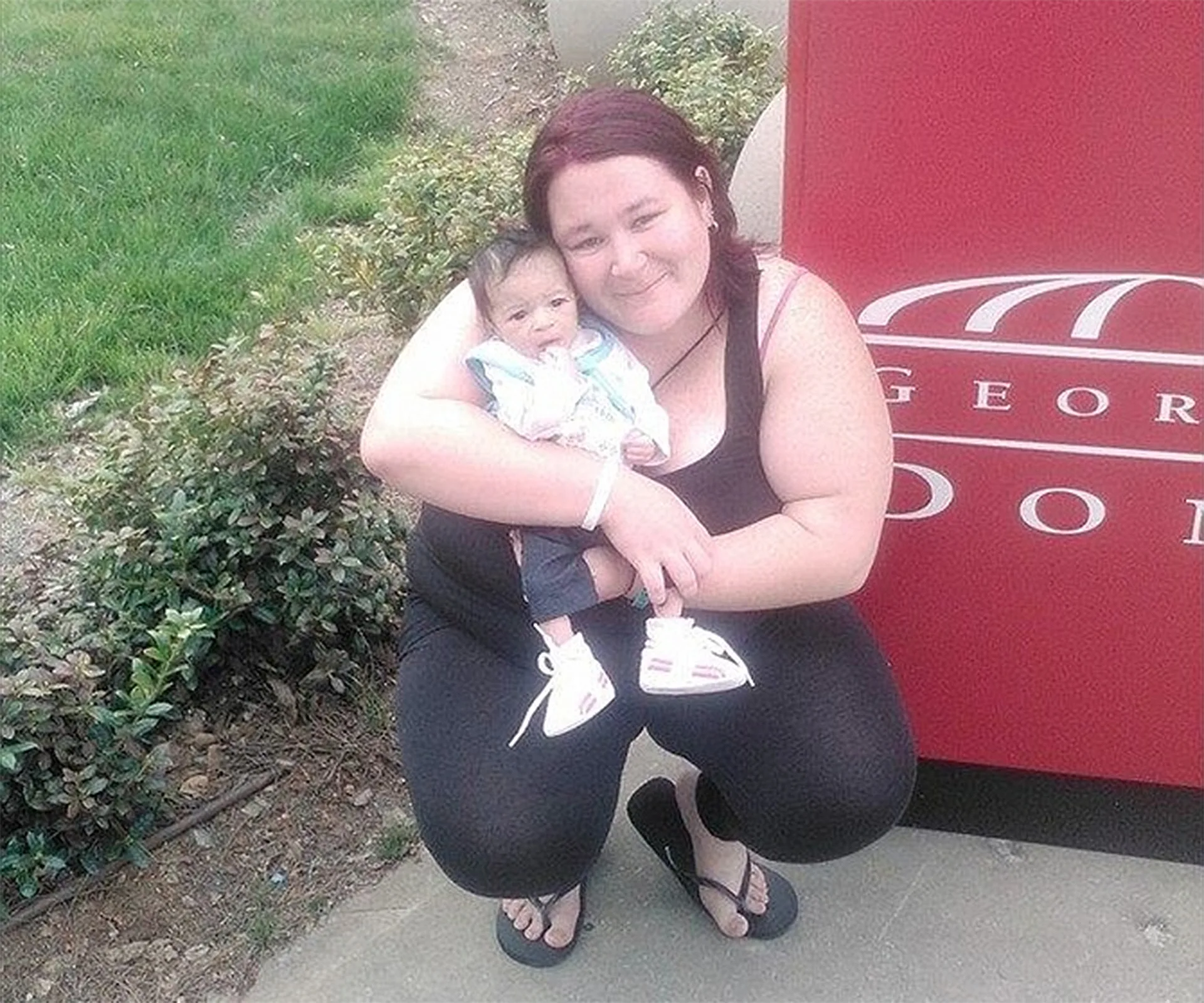 A woman crouches outdoors holding a baby, both smiling near a red sign.