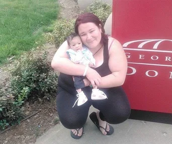 A woman crouches outdoors holding a baby, both smiling near a red sign.