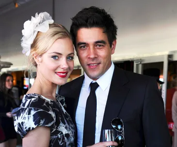 A woman in a floral dress with a white hairpiece and a man in a suit holding a drink, both smiling at an event.