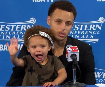Father and young daughter at NBA press conference; daughter waves enthusiastically.