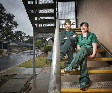 Two people in green prison outfits sit on stairs outdoors in a prison yard, with trees and buildings in the background.