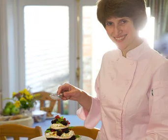 Chef in pink jacket holding a dessert with berries and cream, smiling in a sunlit kitchen setting.