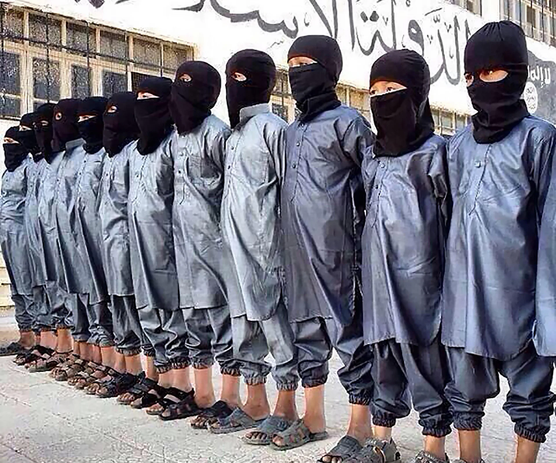 A line of children wearing black uniforms and masks stand against a wall with Arabic writing.